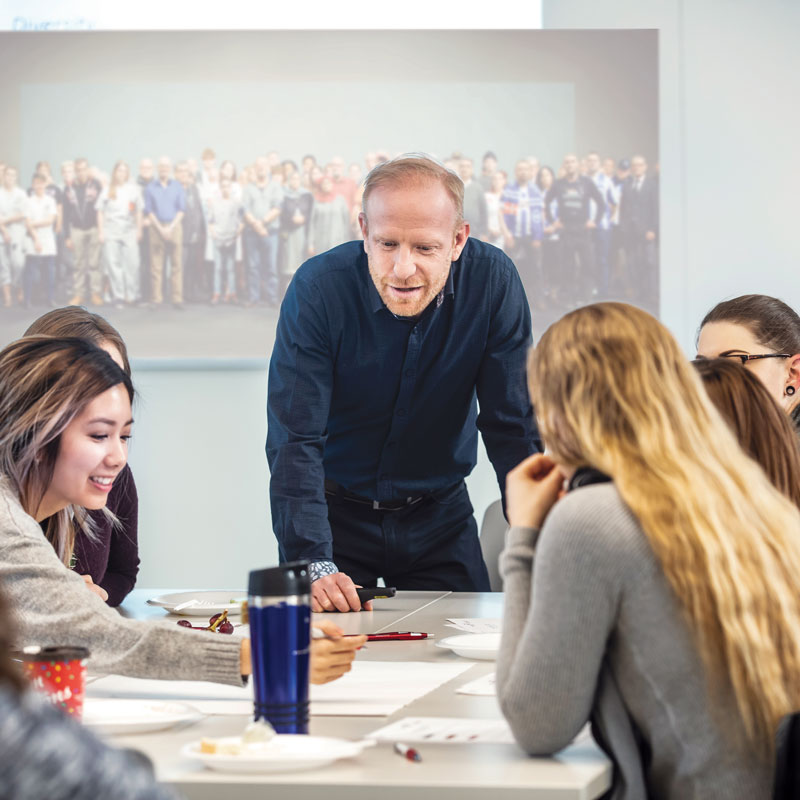 A middle aged man leading leading a group dicussion with several people around a table