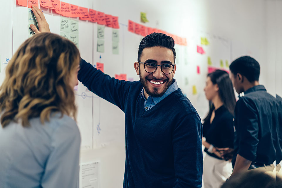 Positive young man laughing while collaborating with colleagues on creating presentation using colorful stickers for productive work in office.Male and female students having fun during workshop