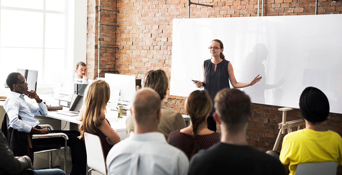 A team of people attending a meeting. A women is in front of a whiteboard presenting.